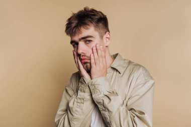 Düşünceli bir ifadesi olan genç bir adam, elleri yüzü üzerinde, bej bir geçmişi var..