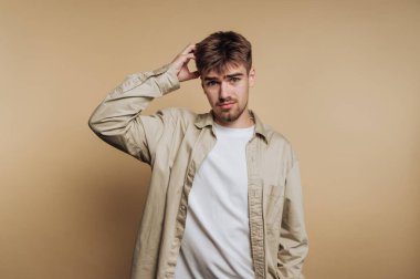 Günlük giysiler içinde başını kaşıyan, bej arka planda genç bir adam..