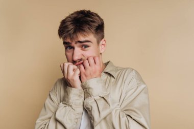 Tırnak yiyen genç bir adam, bej arka planda endişeli görünüyor..