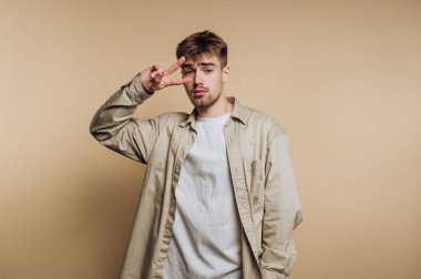 Gündelik kıyafetli genç adam bej arka planda barış işareti yapıyor..