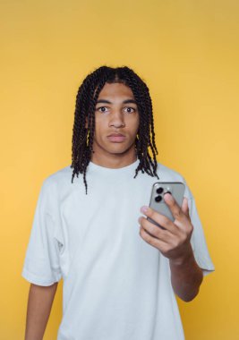 Saçları örgülü, sarı arka planda akıllı telefon tutan genç bir adam..