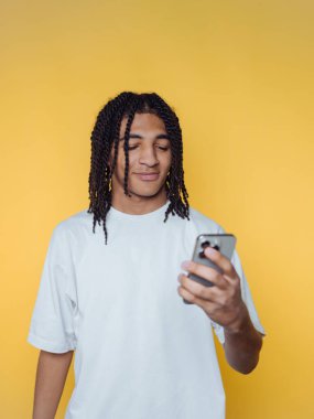 Saçları örgülü genç adam sarı arka planda akıllı telefon kullanıyor..