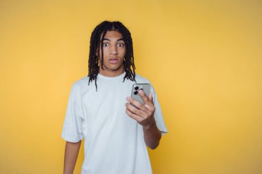 Saçları örgülü, sarı arka planda akıllı telefon tutan şaşırmış genç bir adam..