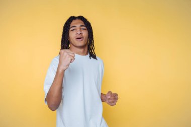 Saçları örgülü genç adam sarı arka planda kutlama yapıyor..