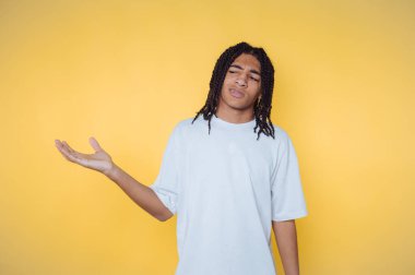 Beyaz tişörtlü, rastalı genç bir adam tek eliyle sarı bir arka plana işaret ediyor..