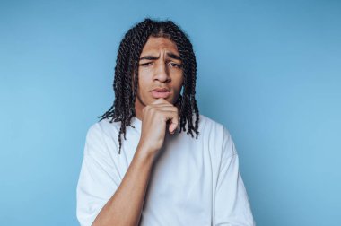 Saçları örgülü genç adam mavi arka planda düşünceli görünüyor..