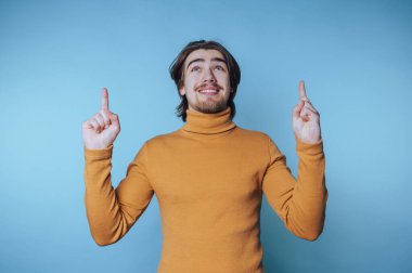 Hardallı süveter giymiş gülümseyen bir adam mavi arka planı işaret ediyor..