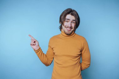 Hardal rengi süveter giyen genç adam yan tarafı işaret ediyor, mavi arka plan..