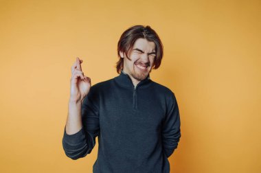 Uzun saçlı genç bir adam, parlak turuncu arka planda parmaklarını çaprazlarken komik bir yüz ifadesi takınıyor.