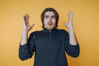 Uzun saçlı ve sakallı, koyu renk bir kazak giyen şaşırmış bir adam, elleri havada sarı bir arka plana karşı duruyor..