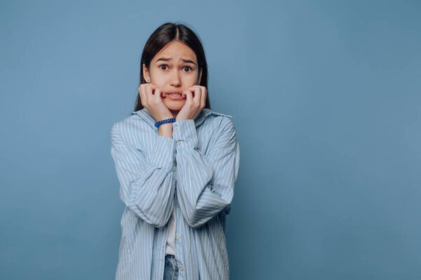 Young woman looking anxious, biting nails, blue background.
