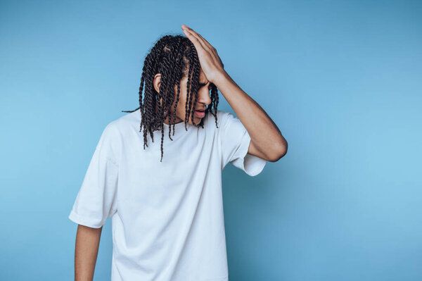 Молодой человек с плетеными волосами в белой рубашке выглядит расстроенным.