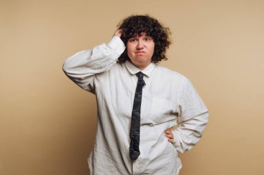 Büyük beyaz gömlek ve siyah kravat takmış, kafası karışmış bir yüz ifadesiyle duran bir insan. Saçları kıvırcık ve kararsız ya da şaşkın görünüyorlar. Arkaplan basit bir bej rengi..