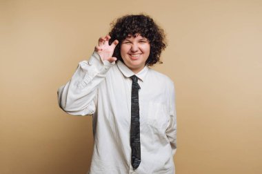 Bir insan bej bir arkaplanda duruyor, gülümsüyor ve tek eliyle bir pençe hareketi yapıyor. Gevşek bir beyaz gömlek ve siyah kravat takıyorlar, neşeli, neşeli bir tavır sergiliyorlar..