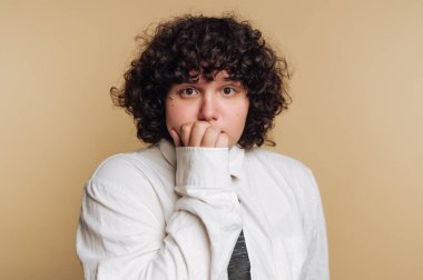 Bir insan bronz bir duvara yaslanır, tırnağını yerken gergin görünür. Kıvırcık saçları var ve beyaz bir gömlek giyiyorlar..