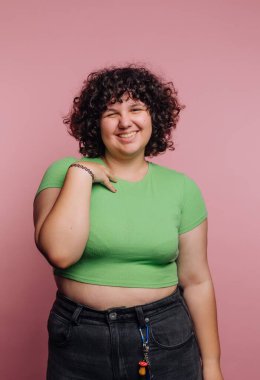 Pembe bir duvarın karşısında duran genç bir kadın parlak bir şekilde gülümsüyor. Yeşil bir bluz giyiyor ve kıvırcık saçları var. Neşeli ifadesi neşeli bir ruh hali sergiliyor..