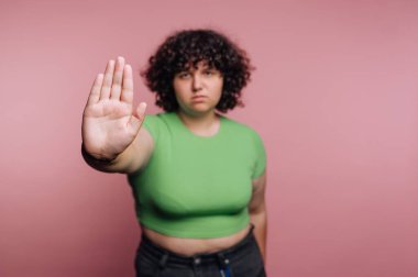 Yumuşak pembe bir zemine karşı bir jest yapmak için elini kaldıran bir insan kendinden emin bir şekilde durur. Sahne, kendini beğenmişlik ve beklentiler üzerine bir mesaj iletiyor..