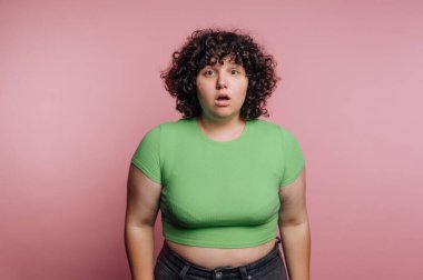 Genç bir kadın pembe bir arkaplana karşı duruyor, şaşırmış bir ifade sergiliyor. Kıvırcık saçları, yeşil bir gömlek ve kot pantolon giyerken yüzünü çerçeveler ve samimi bir anı yakalar..