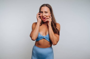 Genç bir kadın hafif bir stüdyoda duruyor, üzerine mavi bir elbise giymiş. Şaşırdığını ve sevindiğini ifade ediyor. Elleri yüzünde. Fotoğraf çekiminde neşeli bir ruh hali sergiliyor..