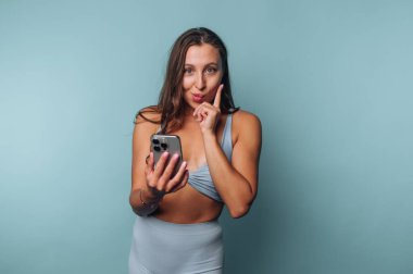 Açık mavi bir duvara karşı poz veren bir kadın akıllı telefonuna bakarken şakacı bir şekilde parmağıyla yanağına dokunuyor. Kendine güveni ve merakı var.