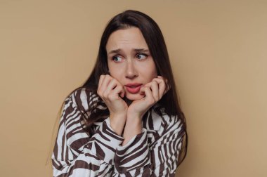 Uzun saçlı bir kadın elleri üzerinde dinlenirken endişeli bir yüz ifadesi gösterir. Arka plan bej renginde, sakin bir atmosfer yaratıyor..