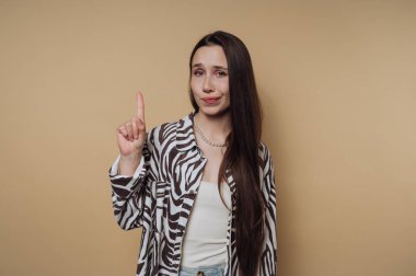 Uzun saçlı genç bir kadın, modaya uygun zebra desenli bir ceket ve sade bir bluz giyer. Gülümsüyor ve parmağını kaldırıyor sanki rahatlamış bir iç mekan ortamında bir noktayı vurgular gibi..
