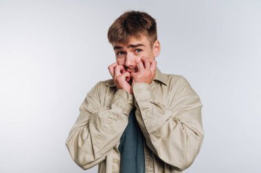 Genç bir adam şaşırmış bir ifade sergiliyor. Elleri ağzını kapatıyor. Endişe ve endişeyi ifade ediyor. Düz bir arkaplanda duruyor..
