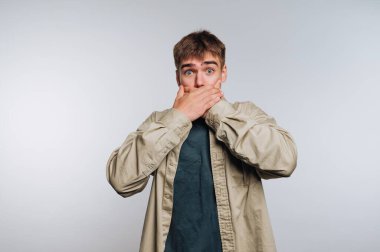 Genç bir adam geniş gözleri ve ağzı açık duruyor, elleriyle kapatıyor, tarafsız bir ortamda şok ya da güvensizlik gösteriyor..