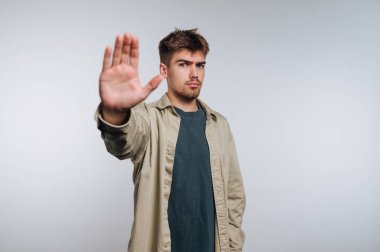 Genç bir adam ciddi bir ifadeyle ayakta duruyor. Avucunu kaldırarak durması için işaret ediyor. Sıradan bir kıyafet giyiyor ve basit bir kapalı mekan ortamında kendinden emin görünüyor..