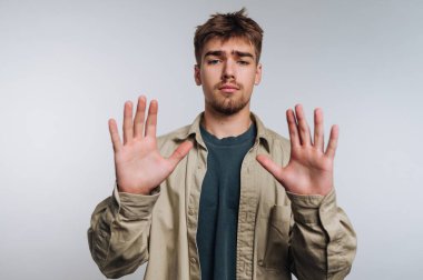 İki eli havada ve avuçları açık genç bir adam ayakta duruyor. Yüzünde ciddi bir ifade var. Açık renkli bir gömlek ve günlük kıyafetler giyiyor. Arka plan basit, nötr bir ton..