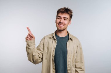Açık bej bir ceketin altında yeşil gömlek giyen genç bir adam gülümsüyor ve sağ işaret parmağıyla açık arkaplanı olan bir stüdyoda yukarı işaret ediyor..
