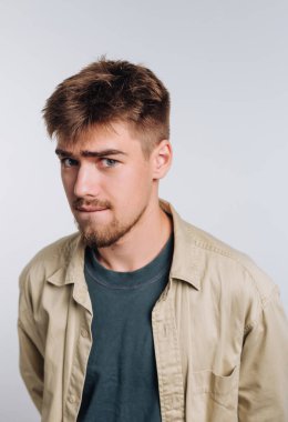 Genç bir adam hafif bir sırıtışla, rahat bir gömlek ve tişört giyerek kendine güveniyor. Kısa saçları ve açık renk sakalı var. Rahat ama çekici bir hava yaratıyor..