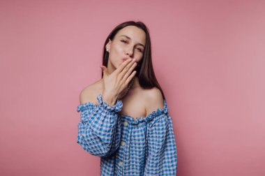 Genç bir kadın pembe bir arka fonun önünde durur ve şakayla bir öpücük savurur. Mavi, kareli, omuzsuz bir bluz giyiyor ve içten gülümsüyor. Kendine güveni ve cazibesi var..