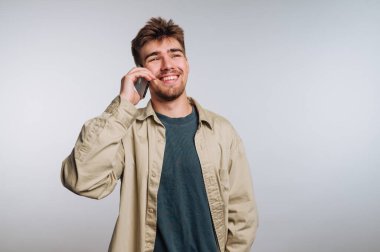 Neşeli genç bir adam gülümsüyor ve parlak bir stüdyoda cep telefonuyla sohbet ediyor. Onun günlük tarzı o anın rahatlamış atmosferine katkı sağlar..