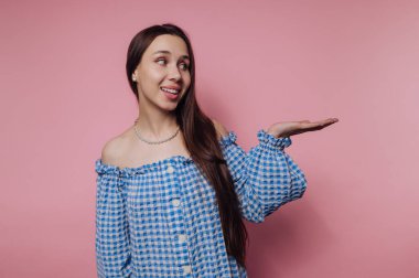 Mavi kareli bluz giyen bir kadın pembe bir duvarın önünde duruyor. Onun uzun saçları ve neşeli bir yüzü var sanki birini davet ediyormuş gibi elini yana doğru uzatıyor..