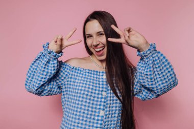 Uzun saçlı, mavi ve beyaz kareli bluz giyen neşeli genç bir kadın ayakta duruyor. Eğlenceli bir fotoğraf çekiminde parlak pembe bir ortamda neşe içinde barış işaretleri gösteriyor..