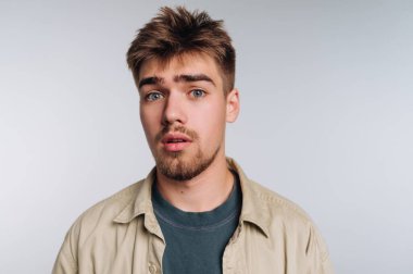 Genç bir adam bir ışık arkaplan karşı duruyor, geniş gözleri ve hafif aralıklı dudakları ile şaşırmış bakıyor. Açık bej bir ceketin altına günlük yeşil bir gömlek giyer..