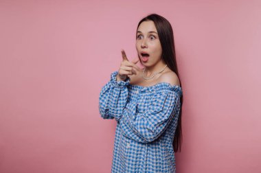 Mavi kareli elbiseli genç bir kadın yüzünde şok olmuş bir ifadeyle dikiliyor. Uzun kahverengi saçları omuzlarının üzerinden akıyor. Yumuşak pembe bir zemine karşı poz verirken..