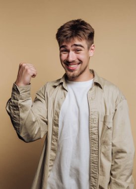 Genç bir adam sevinç anını kutlamak için geniş bir gülümseme sergiliyor. Heyecandan yumruğunu kaldırır, açık renkli bir gömlek giyer ve tarafsız bir arkaplana yaslanır..