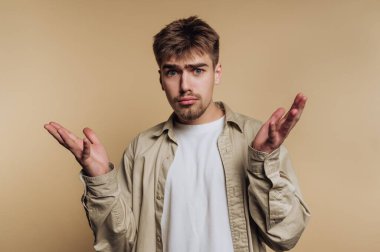 Genç bir adam şaşkın bir bakışla evde duruyor, sorgulayıcı bir şekilde ellerini kaldırıyor. Rahat bir duruşu olan rahat bir kıyafet giyer, hafif bir arkaplanda durur..