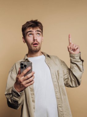 Genç bir adam bir elinde akıllı bir telefon tutarak içeride duruyor. Şaşırmış görünüyor ve diğer eliyle yukarı doğru işaret ediyor. Hafif bir gömlek giymiş..