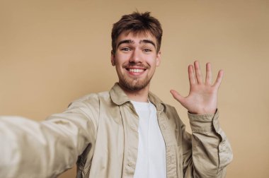Genç bir adam gülümsüyor ve selfie çekerken elini sallıyor. O açık renk bir gömlek giyiyor ve kısa saçlı, basit bir bej duvarın önünde duruyor..