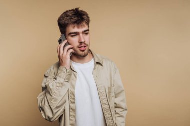 Açık kahverengi saçlı genç bir adam cep telefonuyla bir konuşmayla meşgul. Beyaz bir tişörtün üzerine bej bir gömlek giyer ve basit bir kahverengi fonun karşısında durur..