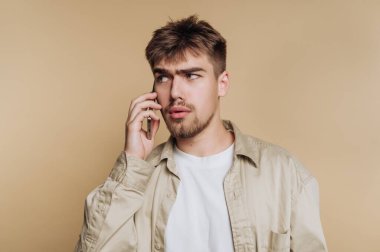 Genç bir adam sıradan bir kıyafet giyerken telefonda konuşuyor. Yumuşak ve tarafsız bir arkaplana karşı konuşmaya odaklanmış görünüyor..