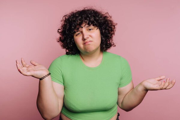 A young person stands with their arms raised in a shrug, displaying a confused expression. They have curly hair and wear a bright green top in front of a soft pink background.