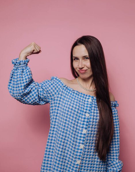 A woman stands against a pink background, flexing her bicep and smiling confidently. She wears a blue checkered shirt and has long, straight hair, radiating empowerment.
