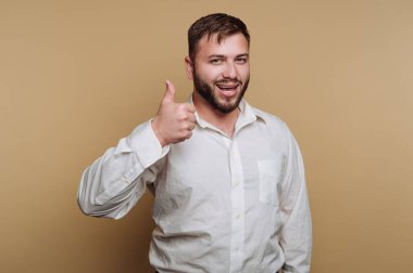 Beyaz gömlekli mutlu bir gencin portresi bej arka planda gülümserken başparmağını kaldırıyor.