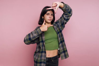 Genç bir kadının portresi ellerini kullanarak yüzünü çerçeveliyor, pembe arka planda sanatsal ifade sergiliyor.