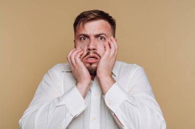 Şaşırmış genç bir adamın portresi ellerini bej arka planda tutarak şoka girdiğini gösteriyor.
