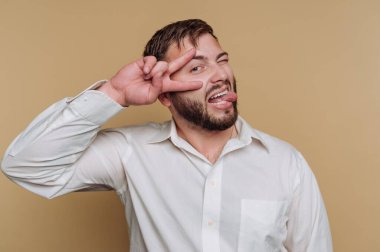 Neşeli genç adamın portresi kameraya komik bir şekilde poz veriyor. Bej arka planda dilini çıkarırken barış işareti yapıyor.